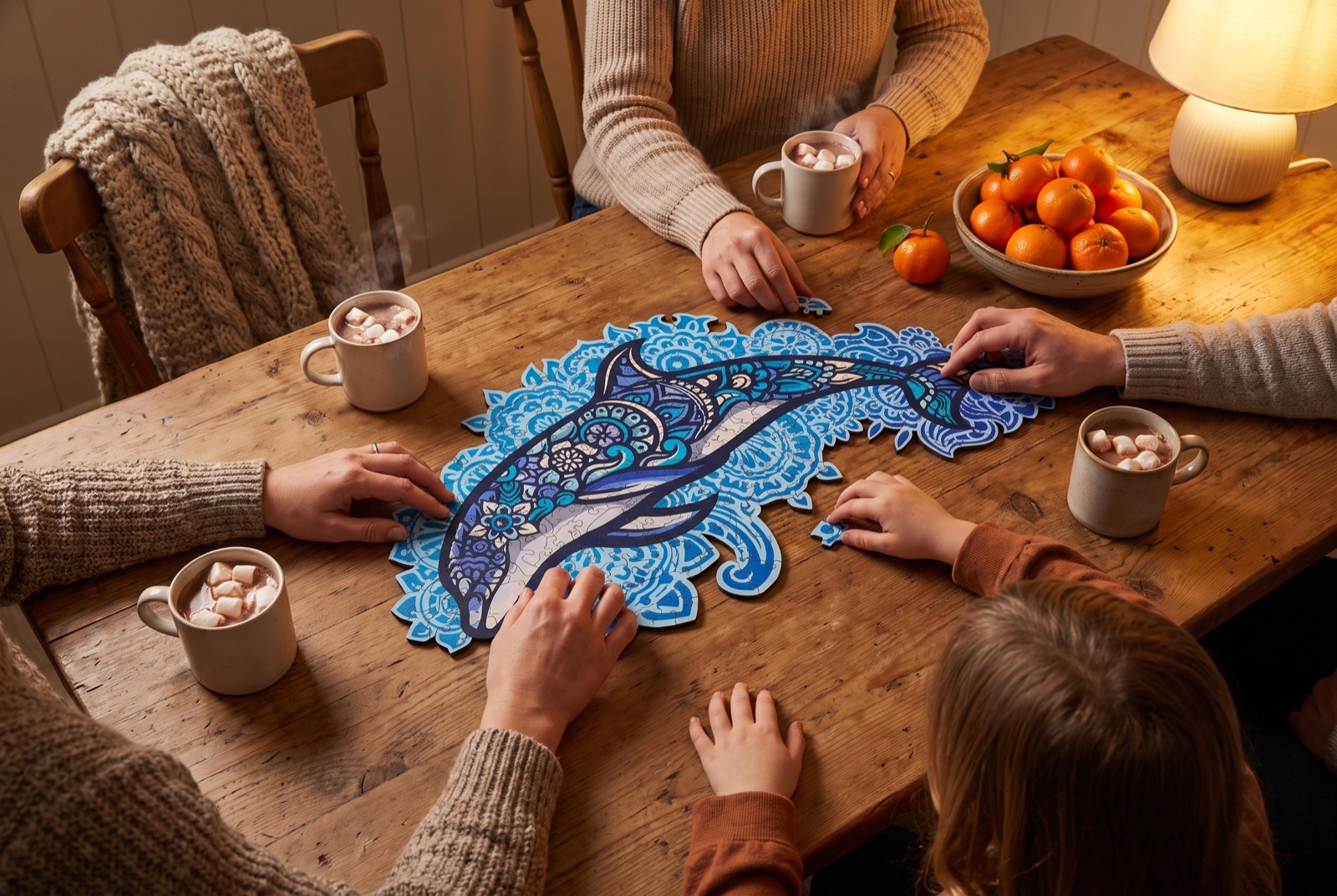 Famille réunie autour d'une table en bois le soir, plusieurs mains adultes et enfants assemblant un puzzle en bois Dauphin Majestueux, avec des tasses de chocolat chaud et un bol de clémentines